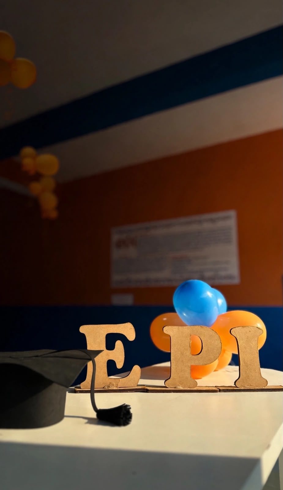 Letras EPI com capelo de formatura e balões azuis e laranja na escola